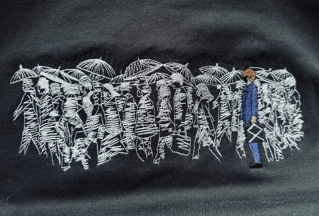 Karoshi embroidery on a black hoodie with workers under umbrellas in white thread.







